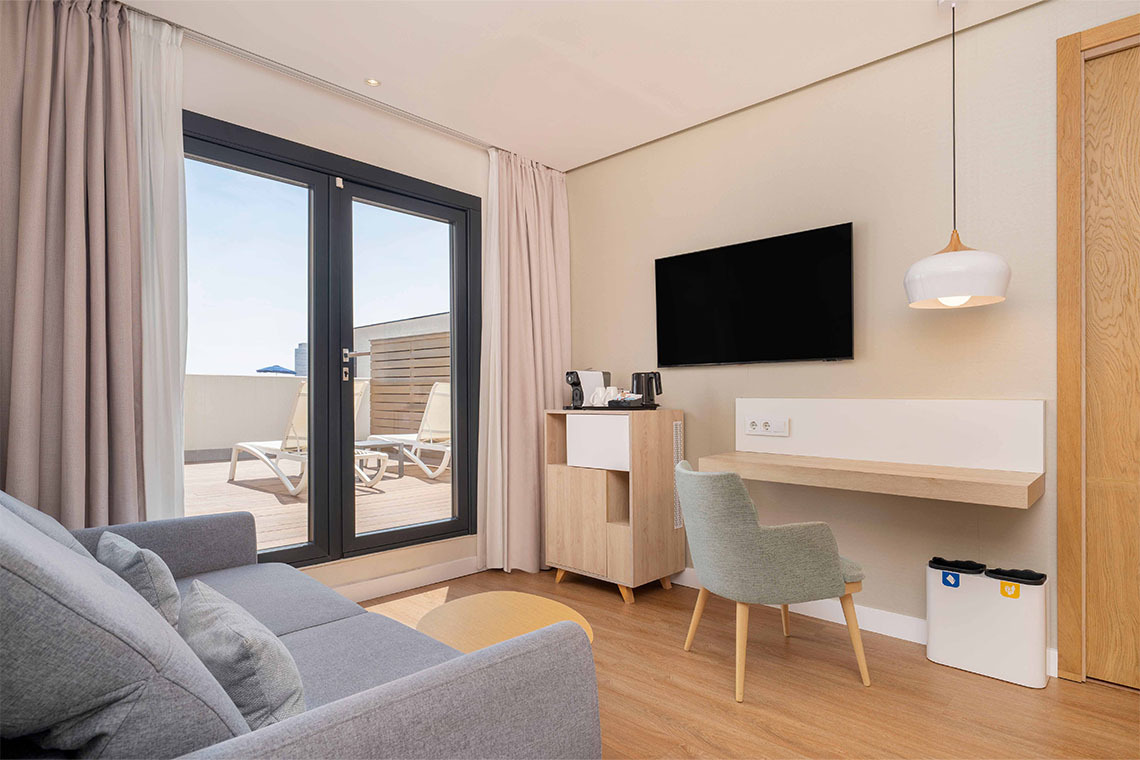 Camera da letto moderna con divano grigio, scrivania e accesso a una terrazza soleggiata.