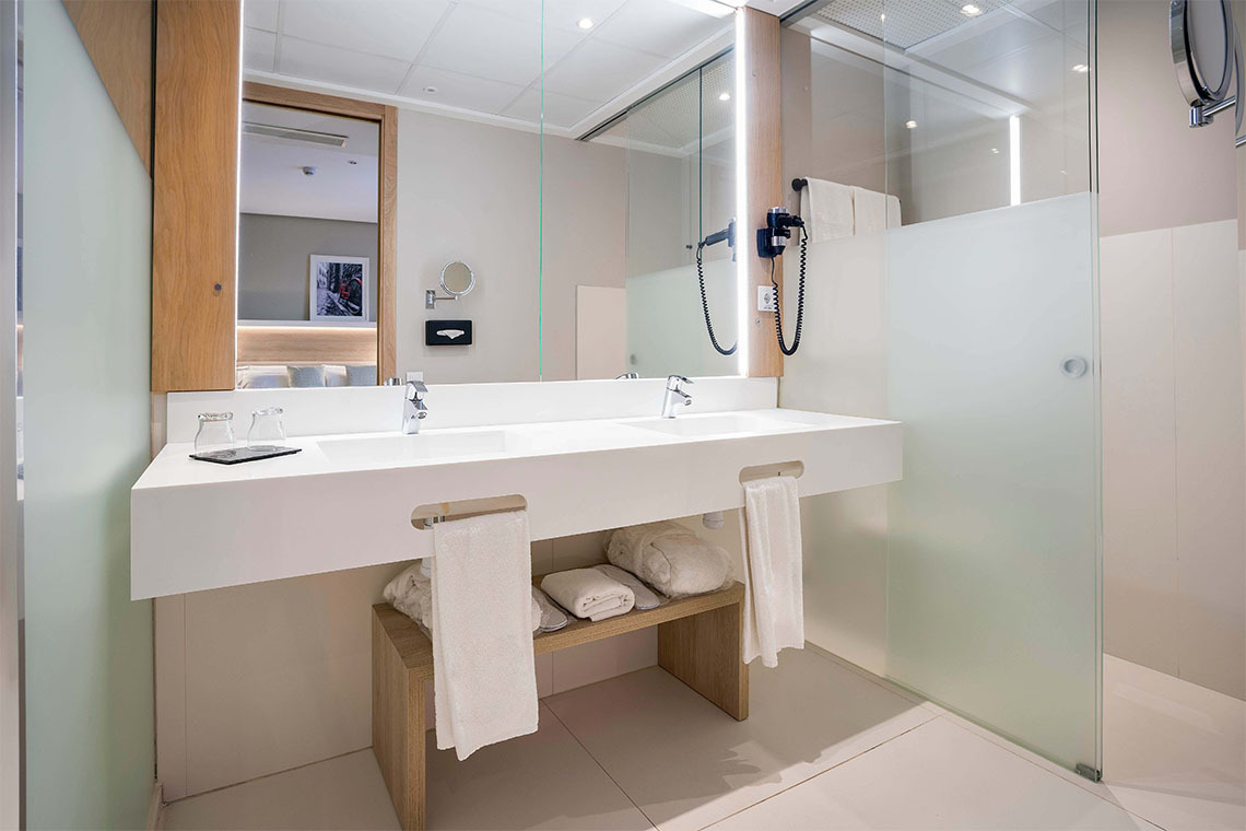 Doble lavabo moderno con toallas blancas, espejo y ducha de cristal en un baño de hotel con ZT: Golden Barcelona.