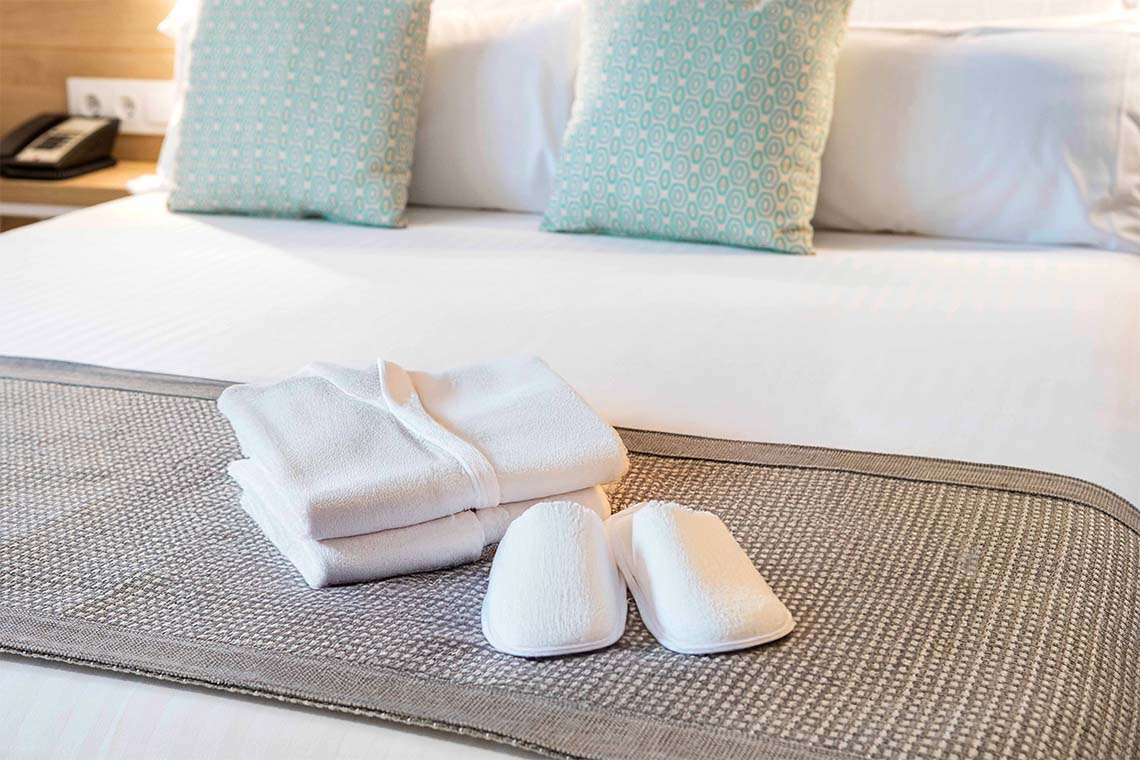Albornoz y zapatillas blancas sobre la cama con cojines turquesas. ZT: Golden Barcelona