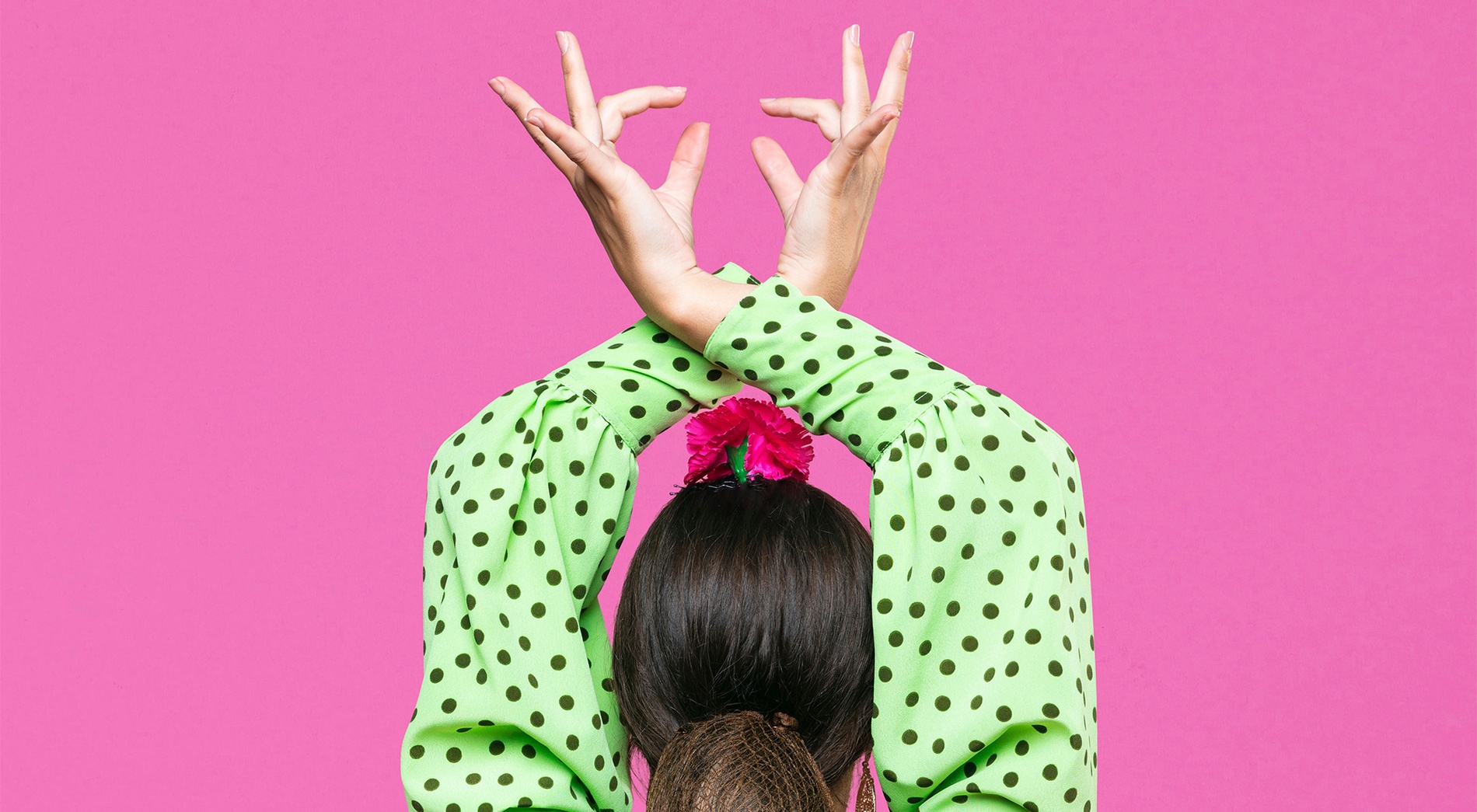A person seen from behind, wearing a green polka-dotted shirt with arms crossed above their head in a dance pose, against a vibrant pink background.