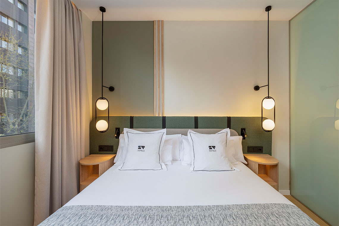 Cama de hotel con lámparas colgantes y almohadas decoradas con el logo ZT: Villa Olimpica.