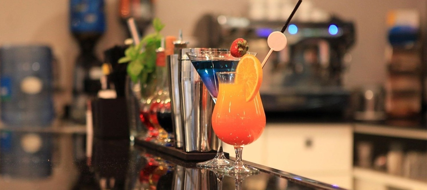 En un mostrador de bar, se ven varios cócteles coloridos y herramientas de bar, reflejándose en una superficie oscura y brillante.